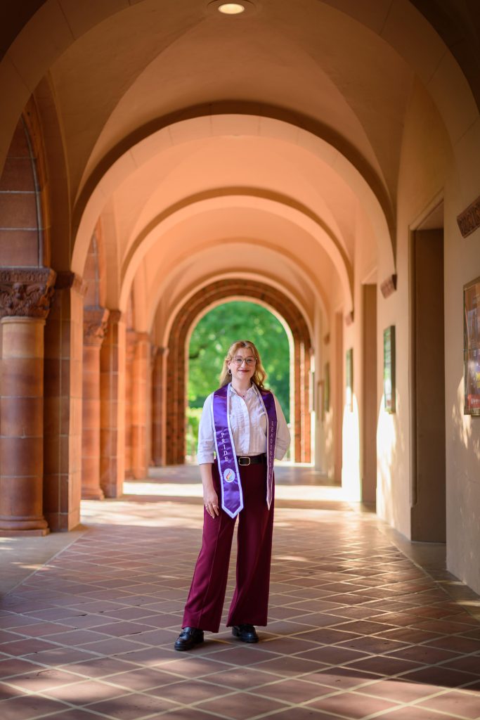 A person wearing a graduation stole stands in the middle of a warmly lit colonnade.