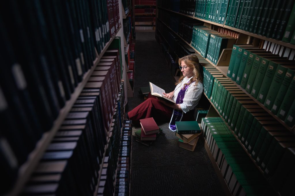 A person wearing a graduation stole sitting in a library surrounded by books is illuminated by a shaft of warm light.